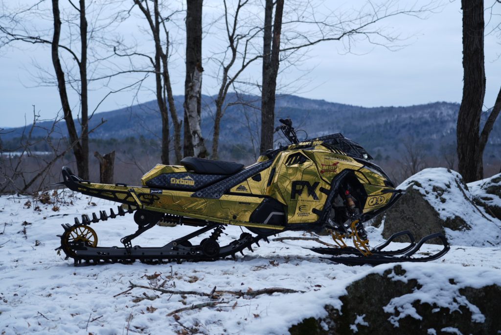 A snowmobile with a custom gold and black wrap parked in a snowy forest with mountain views in the background.