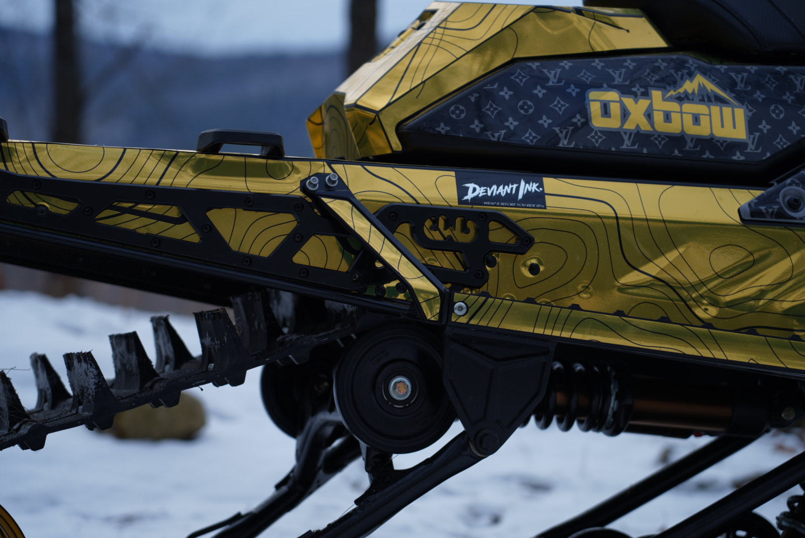 Close-up details of a gold and black snowmobile wrap by Deviant Ink, showing the track, suspension, and Oxbow logo.