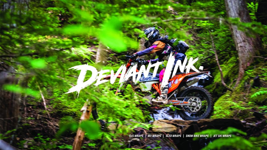 A dirt bike rider in an orange Deviant Ink wrap rides through a forest stream, with the Deviant Ink logo overlaid.