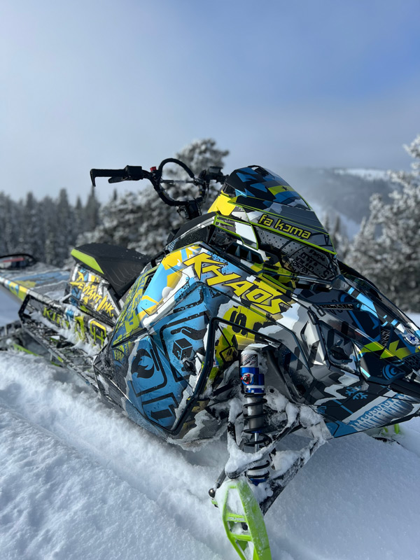 Custom Polaris Khaos snowmobile wrap in electric blue and neon yellow by Deviant Ink, parked in deep powder with mountain backdrop.