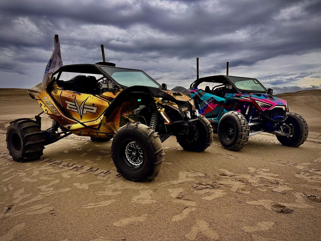 Two Can Am UTVs with custom graphics wraps parked on sandy dunes under a cloudy sky.