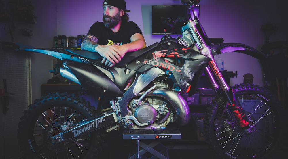 A rider in a garage working on a Honda dirt bike with custom Deviant Ink graphics in black and red.