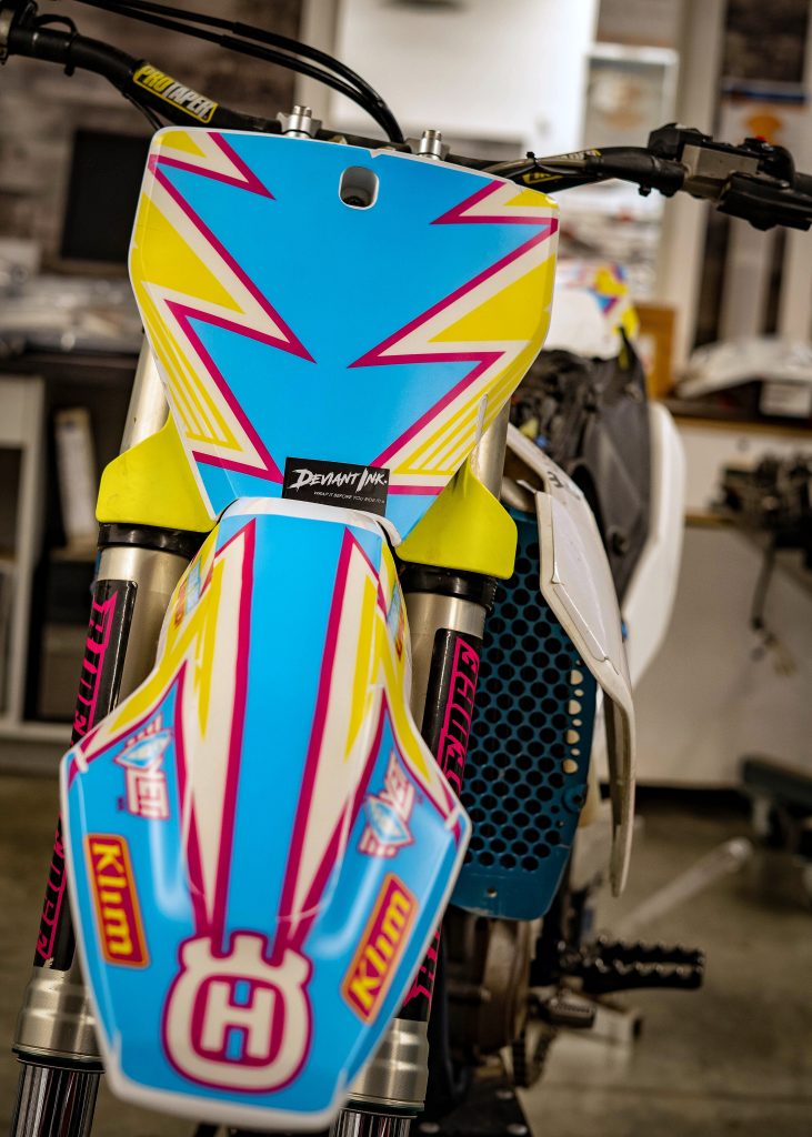 Close-up of a Deviant Ink Husqvarna graphics kit in bright colors, featuring bold yellow and pink accents on a blue background.