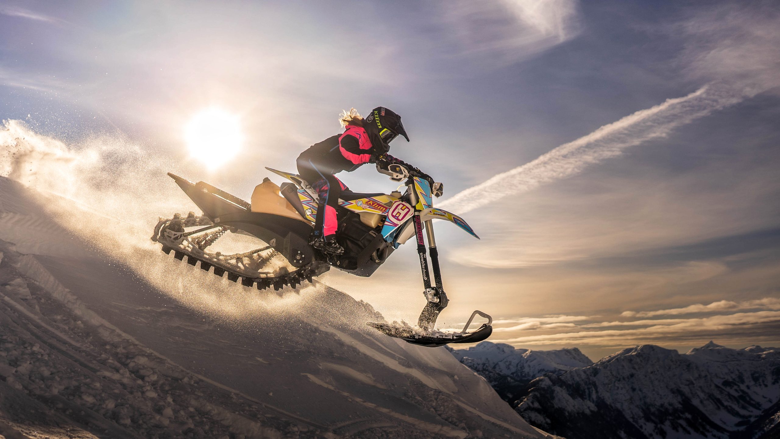 Cass Pro rides a Husqvarna snow bike in custom Deviant Ink graphics, carving powder at sunrise in the mountains.