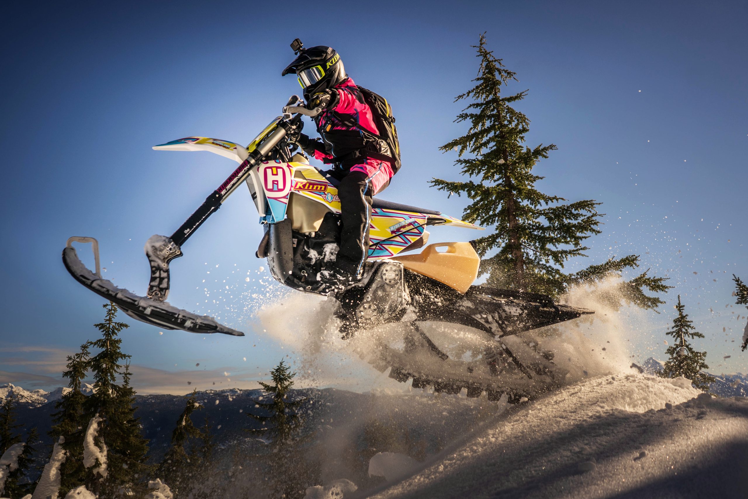 Rider on a Husqvarna snow bike with bright custom graphics, catching air over snowy trees in golden hour light.