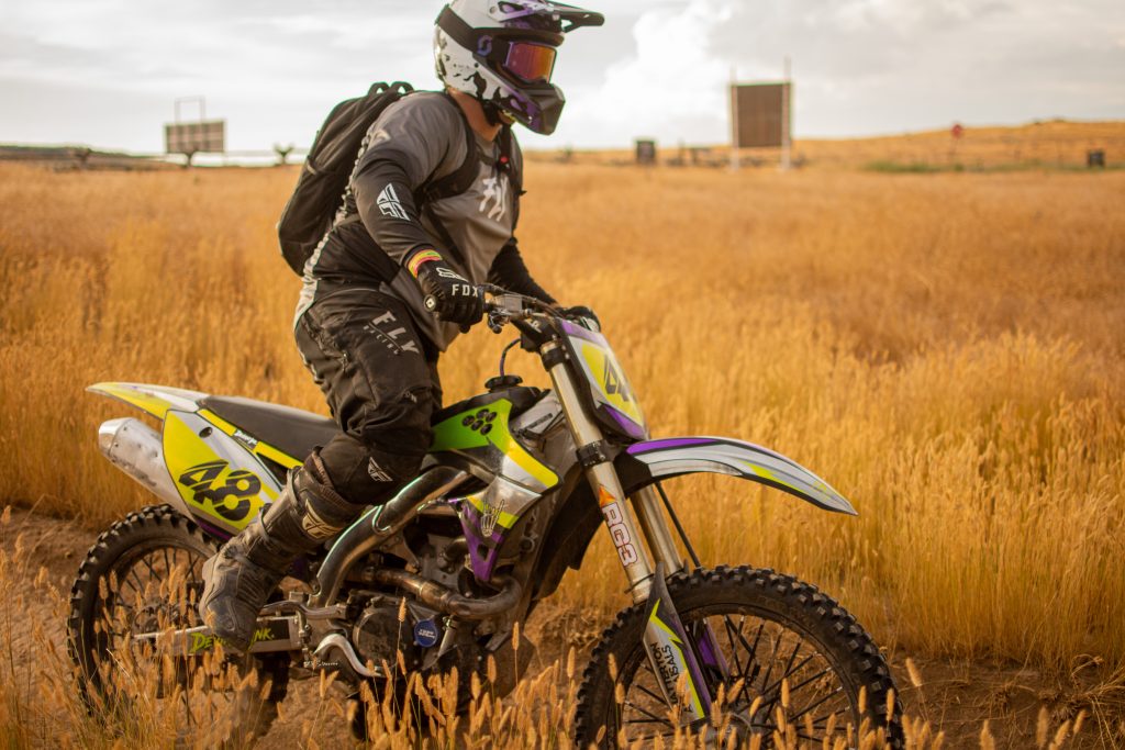 Rider on a dirt bike featuring a custom Deviant Ink sticker set in a yellow, purple, and black color scheme, riding through a golden field.