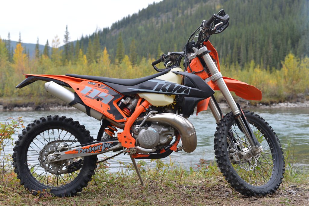 Custom-wrapped KTM dirt bike with Deviant Ink graphics, owned by Janey Rivard, parked near a river in the mountains.