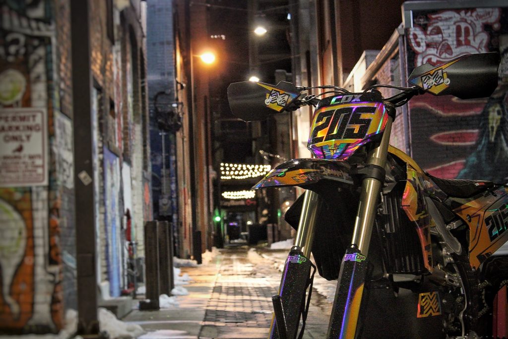 Custom-wrapped dirt bike with holographic graphics by Deviant Ink, photographed in urban alleyway at night