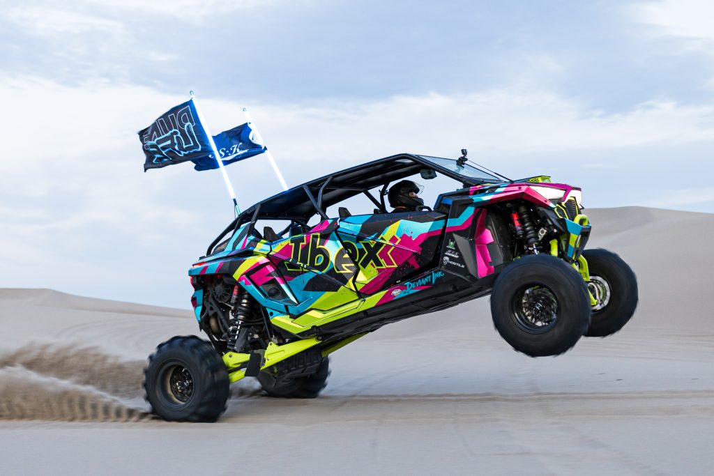 Polaris Pro R flying through sand dunes with neon Deviant Ink wrap and Ibexx branding, lifting off with flags waving